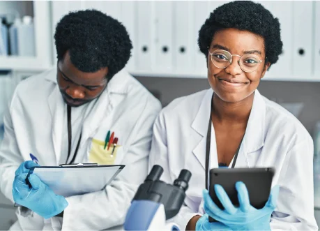 Dois jovens sorrindo enquanto estão estudando juntos utilizando jalecos brancos semelhantes aos de medicos e cientistas