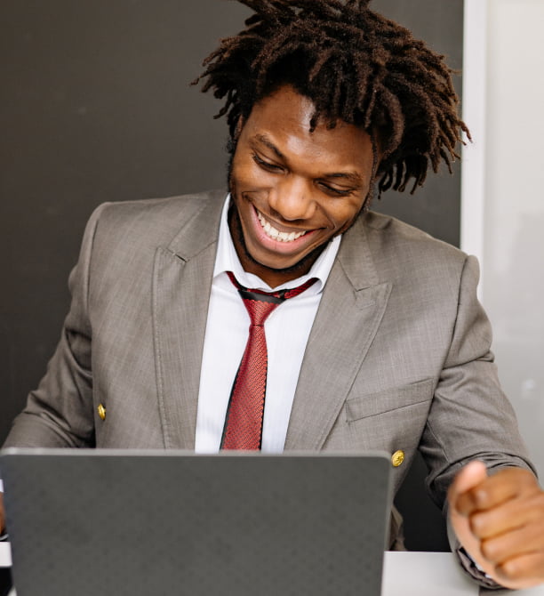 Jovem negro sorrindo utilizando roupa social enquanto está olhando para um notbook
