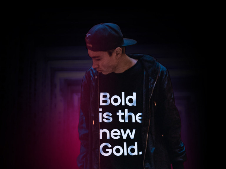 Homem de jaqueta, boné e uma camiseta escrita: Bold is the new gold