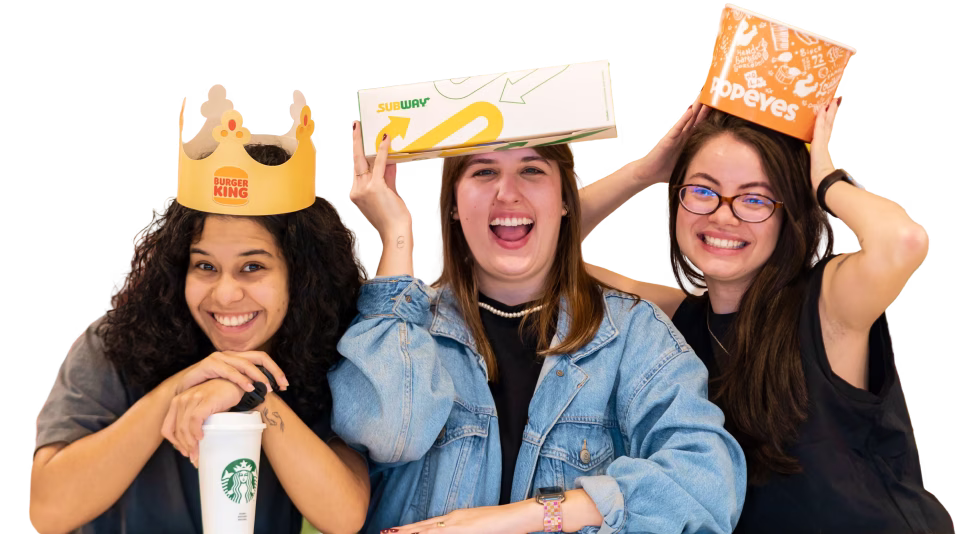 Estagiárias sorrindo segurando produtos do Burguer King, Popeyes, Starbucks e Subway