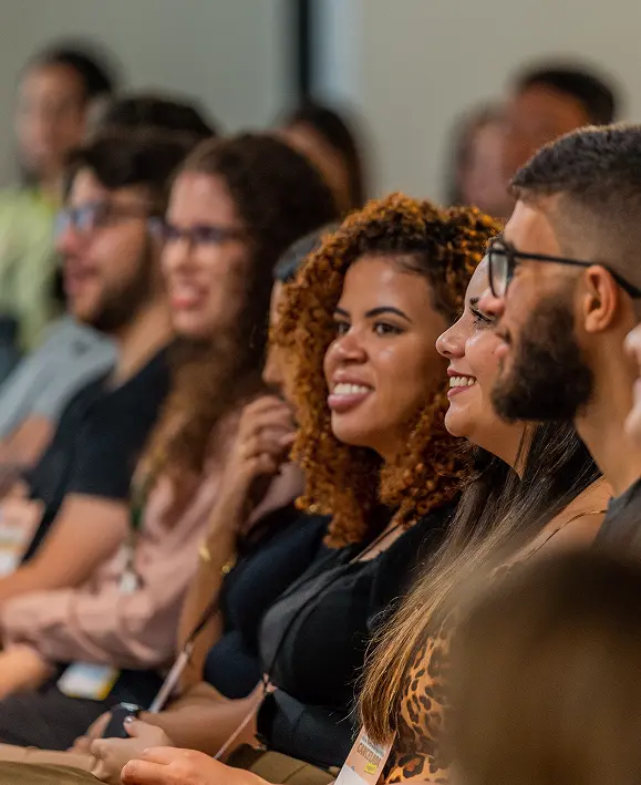 Pessoas na plateia do evento sorrindo e prestando atenção, com foco no rosto de uma mulher negra de cabelos cacheados