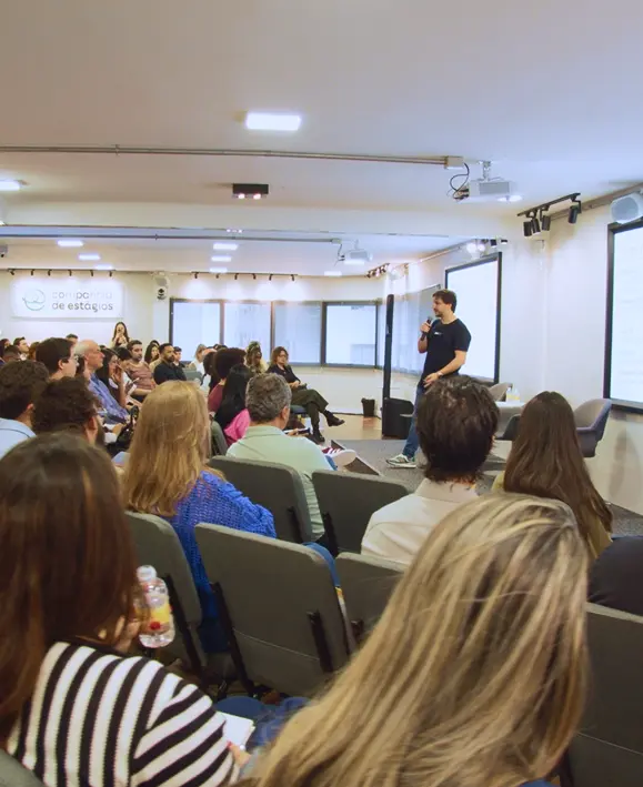 Palestrante fala ao microfone para uma plateia cheia no auditório da Companhia de Estágios. Logotipo da empresa na parede