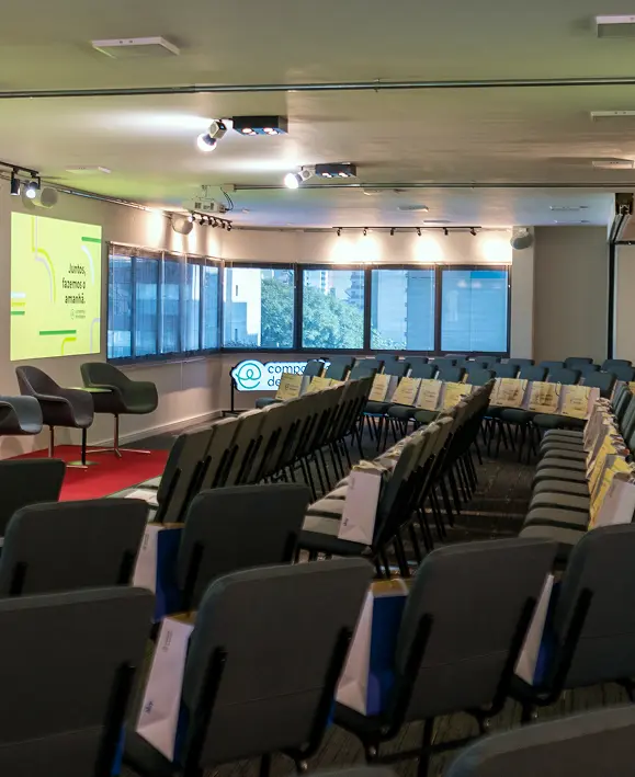 Auditório da Companhia de Estágios vazio, com cadeiras enfileiradas e brindes. Ao fundo, palco com telão exibindo a frase `Juntos, fazemos o amanhã`