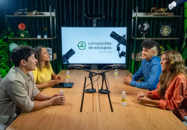 Quatro profissionais gravam conteúdo em formato de mesacast no Estúdio Premium da Companhia de Estágios. Eles estão ao redor de uma mesa com microfones profissionais de ponta, ilustrando a infraestrutura moderna de gravação corporativa oferecida pela marca.